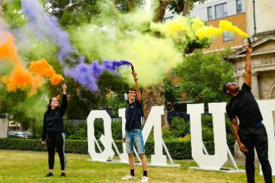 Students holding colourful smoke machines in front of QMUL sign
