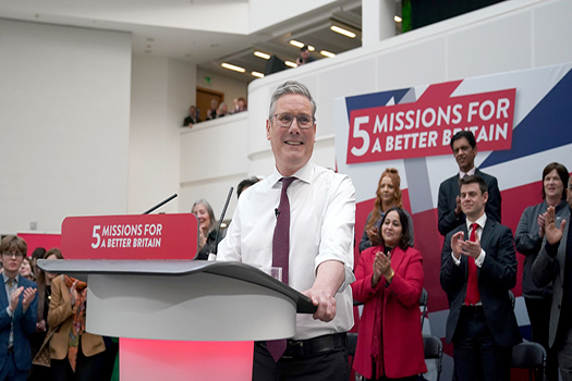 Keir Starmer during one of his 5 Missions speeches. Starmer is wearing a white shirt, red tie, and is standing at a lectern in front of the '5 Missions for a Better Britain' slogan