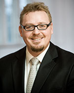 Timo Minssen. He is wearing a black suit jacket, a white shirt and mushroom coloured tie. He is smiling and wearing glasses.