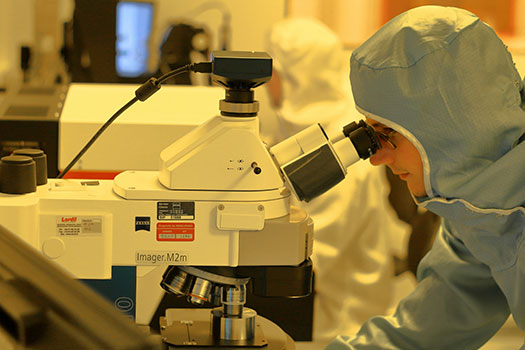 A research looking into an optical microscope. He is wearing a blue lab suit.