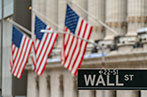 A close up of the Wall Street sign in Manhattan, New York. There are three US flags on flag poles coming off a building in the background.