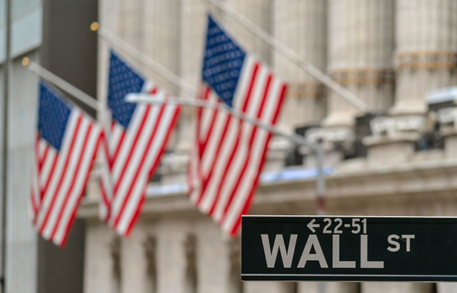 A close up of the Wall Street sign in New York. There are three US flags on flag poles coming off a building in the background.