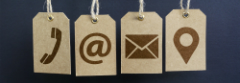 An image of four icons printed on brown paper tags against a dark blue background. The first icon is a telephone handset, the second is an @ symbol, the third is an envelope (email), and the last is a location pin.