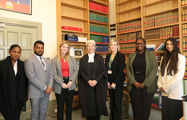 A group photo of Chelsea and Rodney with QMLAC support staff and Right Hon Mrs Justice Stacey standing in from of book shelves.